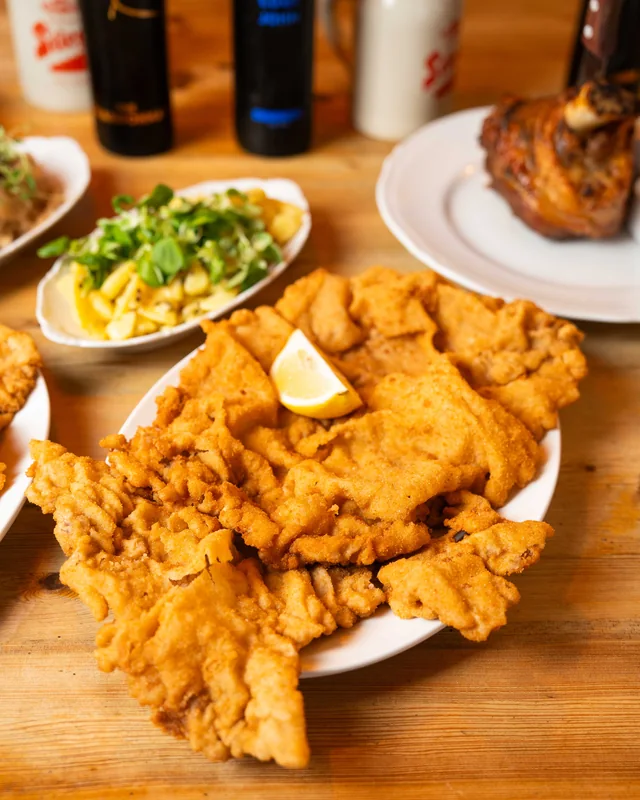 En uppläggning schnitzel med en citronskiva, potatissallad och fläsklägg i bakgrunden
