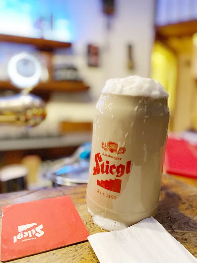 En Mass Stiegl med skum över kanten på ett träbord, på ett rött underlägg