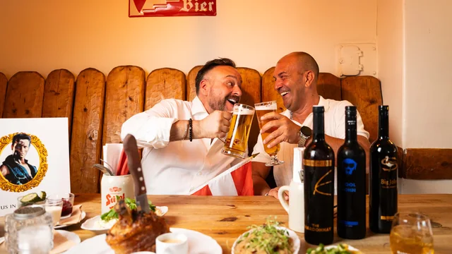 Två män som skålar över en schnitzel och en fläsklägg vid ett dukat bord
