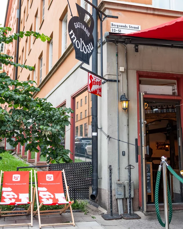 Moldaus entré på Bergsunds Strand med skylten Gasthaus Moldau, röd markis och två Stiegl-solstolar på trottoaren